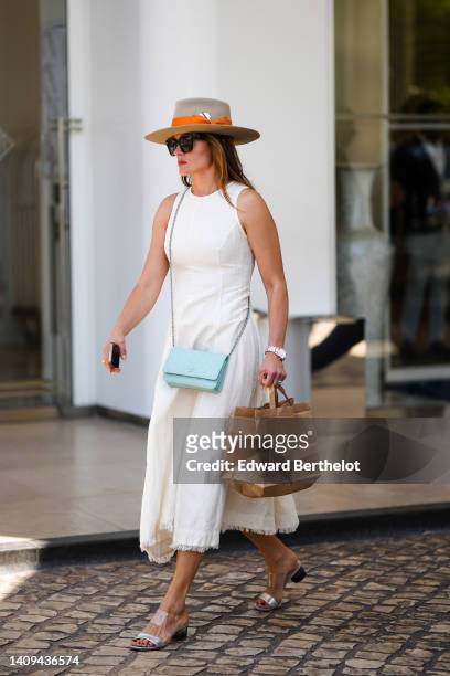 Guest wears a brown felt hat with an orange belt, black sunglasses, a white halter-neck / midi dress, a pale blue leather crossbody WOC from Chanel,...