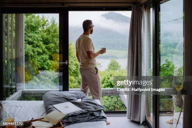 mature man having breakfast and working in tiny house, in front of nature. standing next to window, looking on nature view. - hotel ecologico fotografías e imágenes de stock