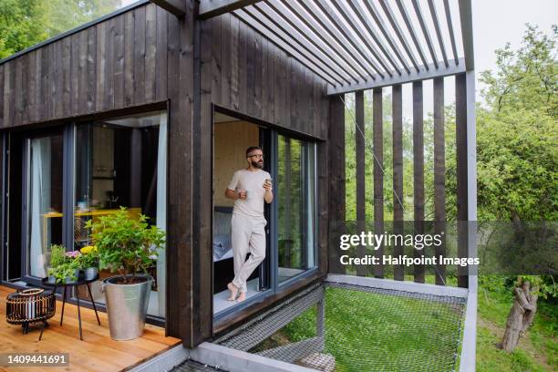 mature man having breakfast and working in tiny house, in front of nature. standing next to window, looking on nature view. - eco house stock pictures, royalty-free photos & images