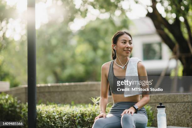 happy young sporty asian woman wear sports outfits with earphone use smartphone listen music sit on the bench and take a rest after workout at home garden park in morning. - woman headphones sport smile iphone stock pictures, royalty-free photos & images