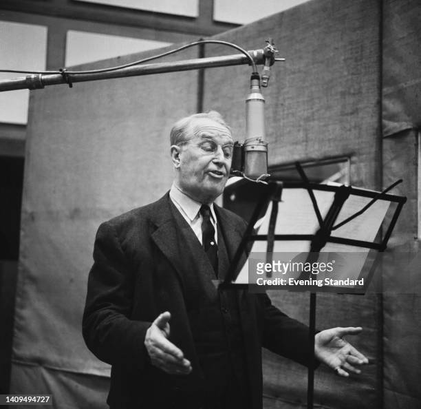 French singer and actor Maurice Chevalier singing into a boom-mounted microphone, the lyrics held on a music stand before him, during an recording...