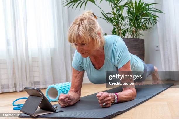 senior womandoing elbow plank on yoga mat in living room - sport tablet stockfoto's en -beelden
