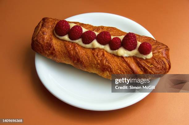 long viennese-style pastry, topped with crème anglaise and fresh raspberries, on a round white plate on an orange cafe table - crème anglaise stock-fotos und bilder