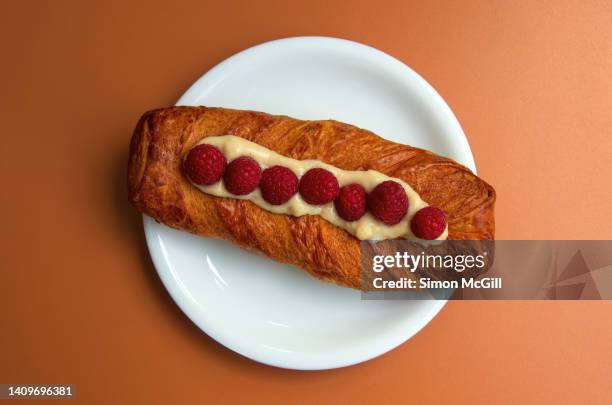 long viennese-style pastry, topped with crème anglaise and fresh raspberries, on a round white plate on an orange cafe table - crème anglaise stock-fotos und bilder