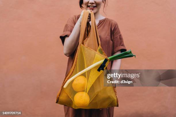 cropped shot of smiling young asian woman carrying a yellow reusable shopping bag, shopping for fresh groceries in the city, standing against orange wall in background. responsible shopping, zero waste, sustainable lifestyle concept - household products stock pictures, royalty-free photos & images