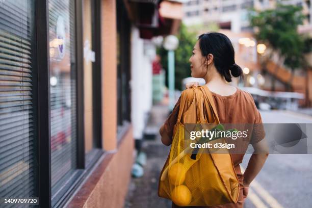 rear view of young asian woman carrying a yellow reusable shopping bag, shopping for fresh fruits and vegetables at supermarket in the city. responsible shopping, zero waste, sustainable lifestyle concept - tote bags photos et images de collection