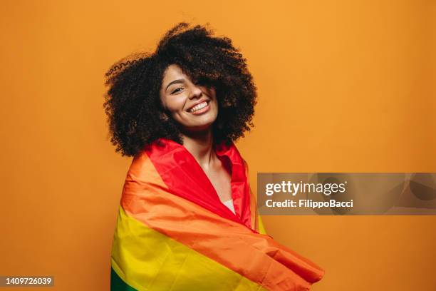 junge erwachsene lächelnde frau vor orangegelbem hintergrund mit regenbogenfahne - regenbogenfahne stock-fotos und bilder