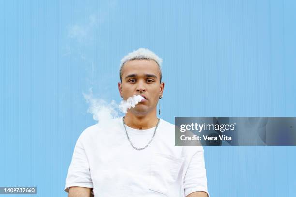 horizontal portrait of skater hispanic man on his 20s smoking outdoors on blue wall - vape stock-fotos und bilder