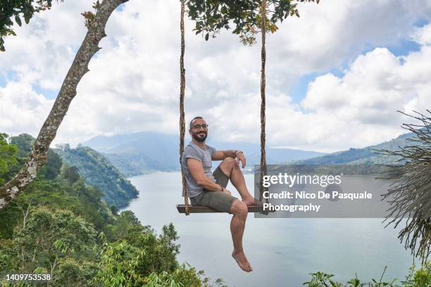 man sitting on swing against lake buyan, bedugul, bali, indonesia - schommelen bungelen stockfoto's en -beelden