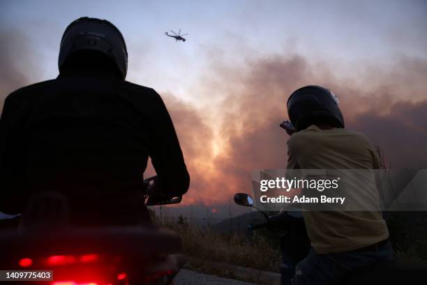 Fire department helicopter passes overhead as motorcyclists look at wildfires burning a residential area just outside Athens on the slopes of the...