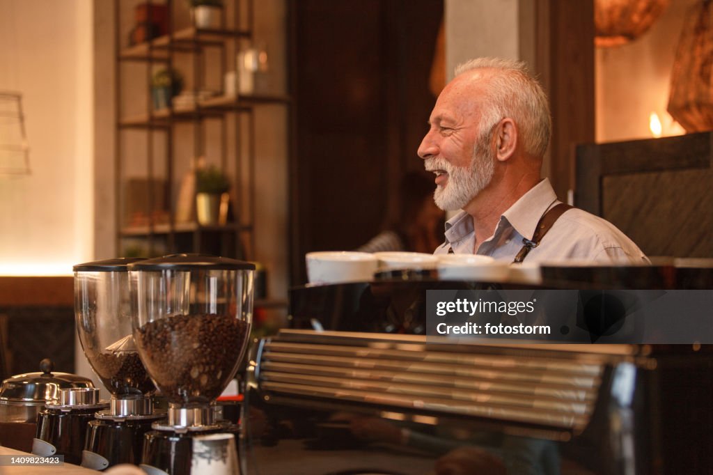 Charming senior barista making drinks behind bar counter and chatting with customers