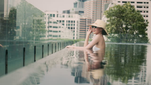 Smiling Asian female wearing hat and sunglass stay in pool at hotel.