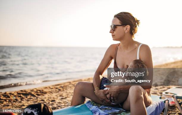 mujer sentada en la playa y amamantando a su bebé - pechos de mujer playa fotografías e imágenes de stock