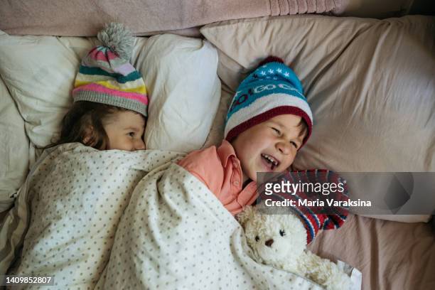 closeup of a laughing boy with his sister laying in their bed wearing winter hats - energieeffizienz stock-fotos und bilder