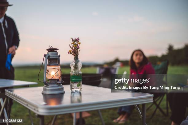 close up of oil lamp & flowers in diy vase on camping table with couple enjoying the sunset view at camping field in background - garden table stock-fotos und bilder