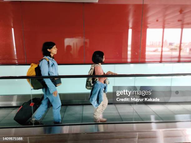 mother and daughter on a moving walkway at the airport carrying their luggage - cinta mecánica fotografías e imágenes de stock