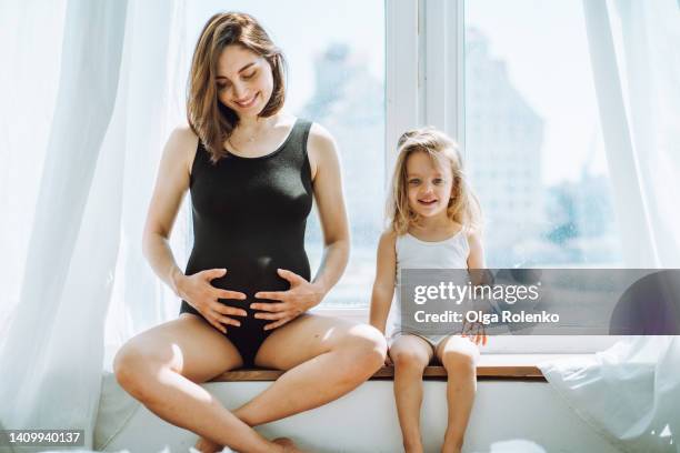 smiling pregnant dark-haired woman touching her belly and little girl sit on white windowsill in curtains - child white stock pictures, royalty-free photos & images