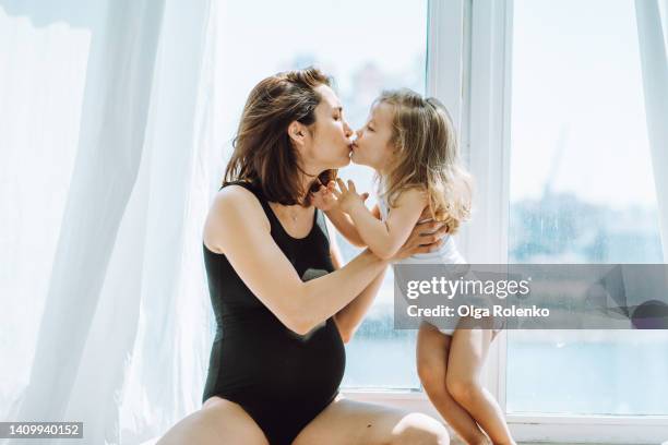 pregnant dark-haired woman kissing her little daughter, sitting by the white window - parents baby sister stock pictures, royalty-free photos & images