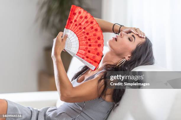 woman suffering a heat wave using a fan, lying on a couch in the living room at home. - ondata di calore foto e immagini stock