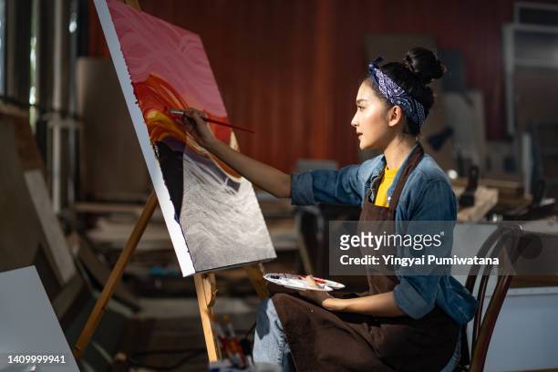 smiling portrait on young asia female artist,she sitting in front of her big painting on canvas.asian woman spending weekend morning painting in studio room at home - pittore artista foto e immagini stock