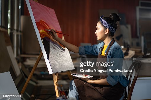 Smiling portrait on young asia female artist,she sitting in front of her big painting on canvas.Asian woman spending weekend morning painting in studio room at home Smiling portrait on young asia female artist,she sitting in front of her big painting on canvas.Asian woman spending weekend morning painting in studio room at home