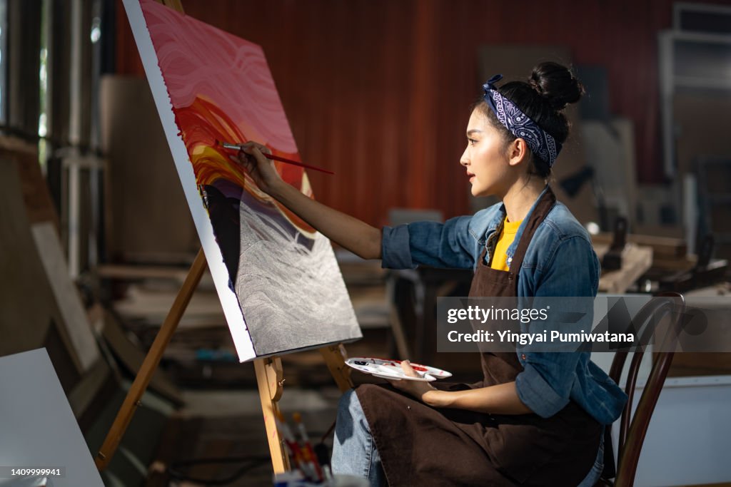Smiling portrait on young asia female artist,she sitting in front of her big painting on canvas.Asian woman spending weekend morning painting in studio room at home