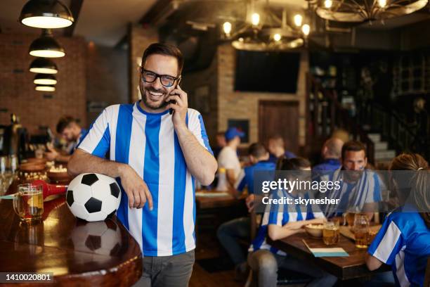 happy soccer fan talking on mobile phone in a full pub. - football phone stock pictures, royalty-free photos & images