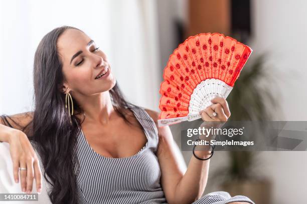 woman suffering a heat wave using a fan, sitting on a couch in the living room at home. - ac weary stock pictures, royalty-free photos & images