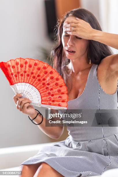 woman suffering a heat wave using a fan, sitting on a couch in the living room at home. - ac weary stockfoto's en -beelden