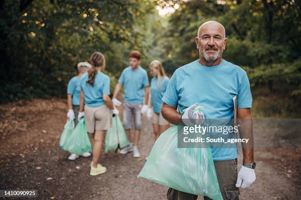 portrait of senior volunteer while cleaning garbage with group of volunteers in nature - garbage man stockfoto's en -beelden