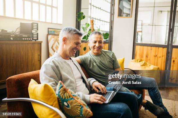 gay couple on couch  going over their home finances - mature adult on laptop stock pictures, royalty-free photos & images