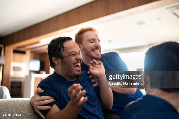 young man with special needs celebrating with friends while watching sports match at home - social tv awards stockfoto's en -beelden