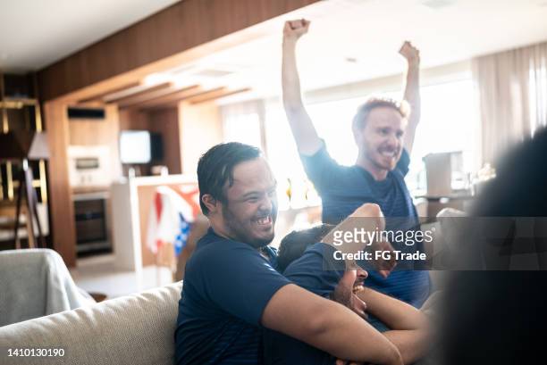 young man with special needs celebrating with friends while watching sports match at home - social tv awards stockfoto's en -beelden