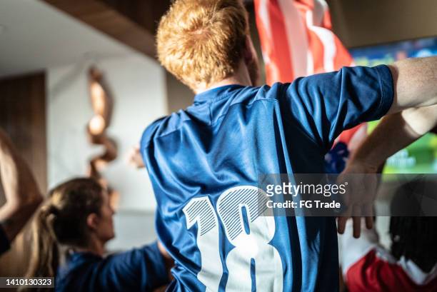 rear view of a sports fan celebrating holding american flag at home - camisola de futebol imagens e fotografias de stock