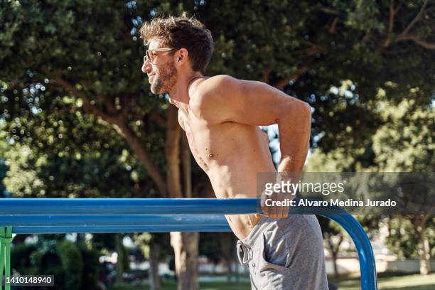 side view of a young muscular man training dips on a gymnastic parallel bars in a public park. - parallel bars gymnastics equipment stock-fotos und bilder
