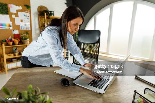 jovem empresária em pé sobre sua mesa, verificando algo no laptop rapidamente - terminar sessão - fotografias e filmes do acervo