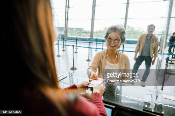 sorrindo viajante asiática asiática sênior em pé em um balcão de check-in do aeroporto e esperando sua passagem aérea e cartão de embarque - passagem de avião - fotografias e filmes do acervo