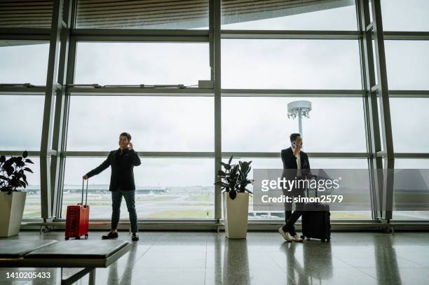 shot of full length of two asian chinese businessmen using smartphone while standing through window of an airport terminal. - kuala lumpur airport stock pictures, royalty-free photos & images