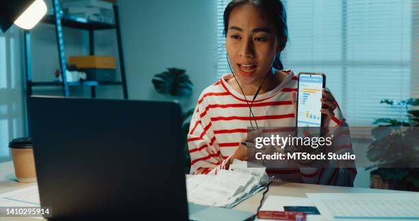 jovem asiática planejadora financeira mulher com suéter usar fone de ouvido com laptop on-line reunião presente utilitário de despesas mensais, cartão de crédito plano financeiro mensal em smartphone em casa à noite. - pointing at camera - fotografias e filmes do acervo