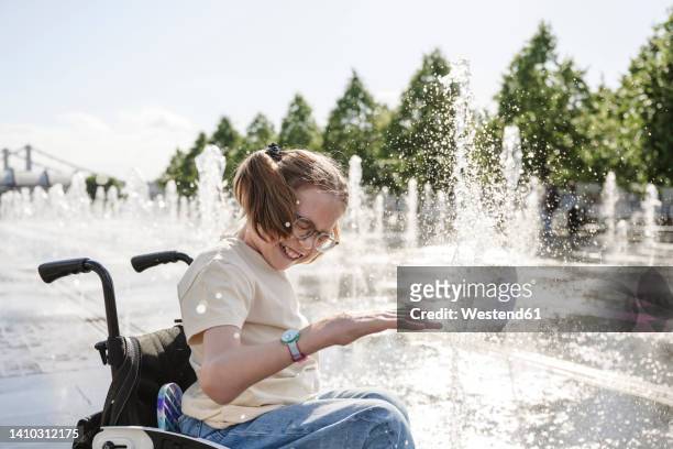 cheerful girl with disability sitting on wheelchair playing by fountain - körperliche behinderung stock-fotos und bilder