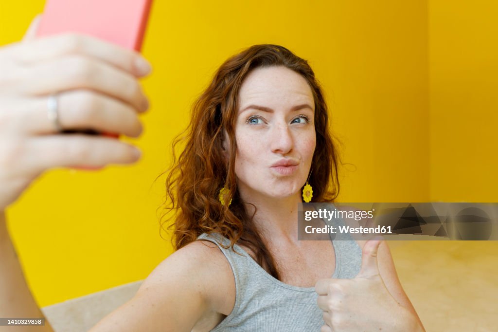 Woman gesturing thumbs up taking selfie through smart phone in front of yellow wall