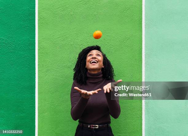 cheerful woman with mouth open throwing orange in front of green wall - catch stockfoto's en -beelden