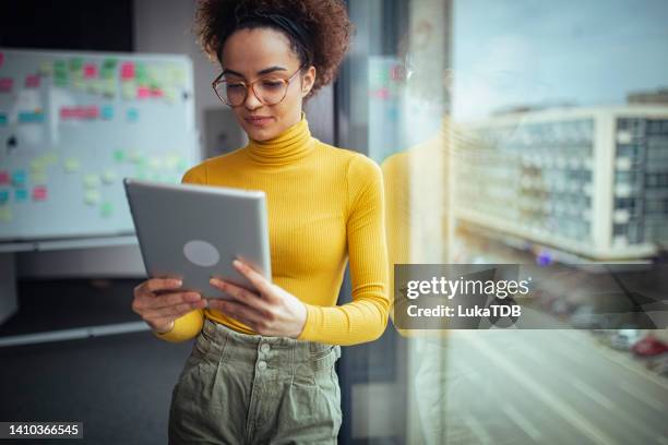 una giovane donna d'affari usa un tablet mentre si trova in un ufficio vicino alla finestra. - yellow blouse foto e immagini stock