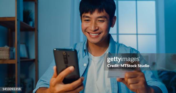 nahaufnahme eines jungen mannes, der telefon und kreditkarte für online-einkäufe benutzt, sitzt nachts am schreibtisch im wohnzimmer zu hause. - phone credit card stock-fotos und bilder