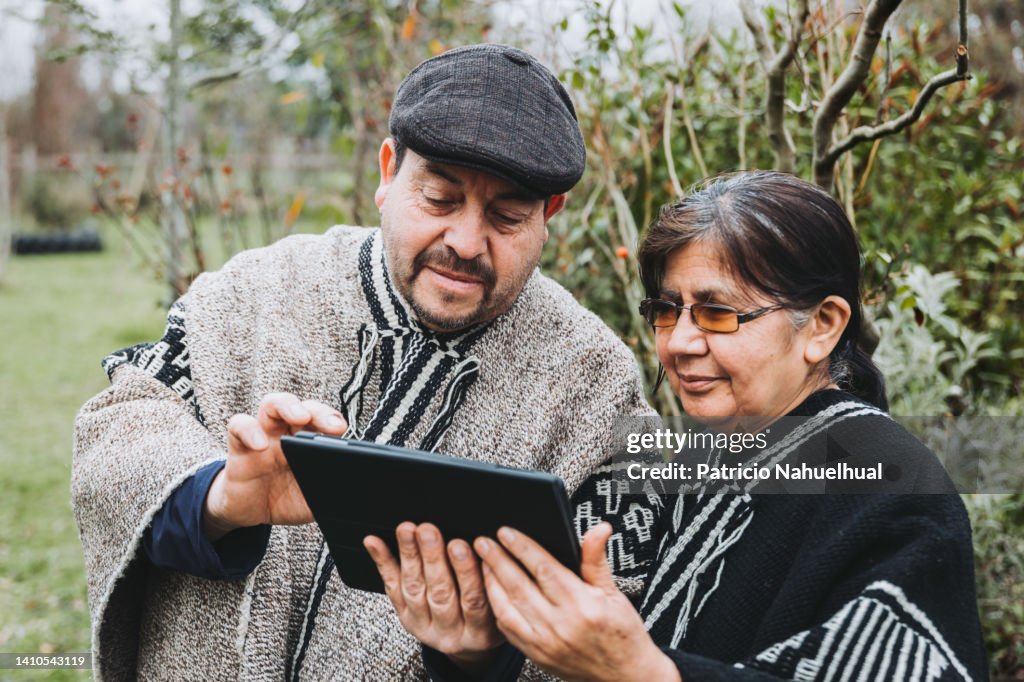Two smiling traditional mapuche people dressed in gray and black wool ponchos, using a tablet to make a video call