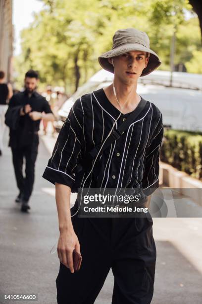 Model wears a tan bucket hat, black striped baseball-style button up shirt, brown belt, black pants, white sneakers and wired headphones after the...