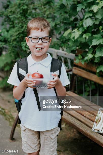 a schoolboy in glasses 6-7 years old in a white shirt and shorts with a backpack and a lunchbox in his hands, eats a cucumber while standing on the street - 8 9 years stock pictures, royalty-free photos & images