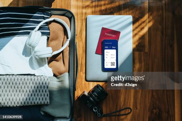 flat lay shot of open suitcase packed with clothings, headphones, camera, laptop and passport on wooden floor against sunlight, with smartphone device screen showing electronic flight ticket itinerary. travellers accessories. travel and vacation concept - plane ticket stock pictures, royalty-free photos & images