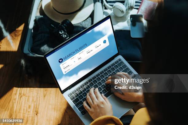 young asian woman shopping online for flight ticket on airline website with laptop at home, sitting next to suitcase with sun hat, camera, clothings, headphones, smartphone and passport. getting ready for vacation. travel and vacation concept - reserveren stockfoto's en -beelden