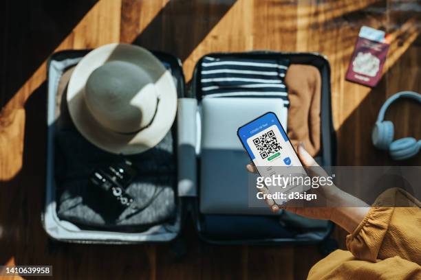cropped shot of woman's hand holding smartphone showing digital certificate of covid-19 vaccination above an open suitcase with clothings, camera, hat, laptop, passport and headphones on wooden floor. traveller's accessories. travel and vacation concept - phone screen at airport stockfoto's en -beelden